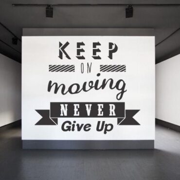 Primer izgleda črne samolepilne stenske nalepke Keep on moving never give up na beli steni. Nalepka je napis, ki se glasi: Keep on moving never give up.