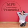 Primer izgleda temno rdeče samolepilne stenske nalepke Life is about na rdeči steni. Nalepka je napis, ki se glasi: Life isn't about finding yourself. LIFE is about creating yourself.