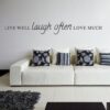 Primer izgleda črne samolepilne stenske nalepke Live well Laugh often Love much na beli steni v dnevni sobi. Nalepka je napis, ki se glasi: Live well, Laugh often, Love much.