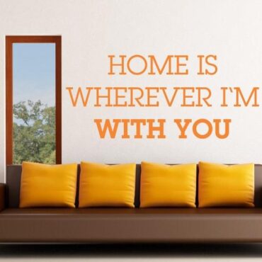 Primer izgleda oranžne samolepilne stenske nalepke Home is wherever Im with you na bež steni v dnevni sobi.