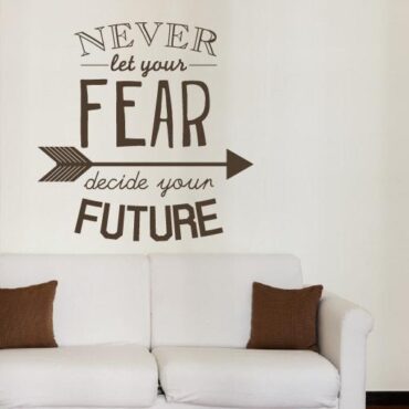 Primer izgleda čokoladno rjave samolepilne stenske nalepke Never let your fear decide your future na beli steni v dnevni sobi. Nalepka je napis, ki se glasi: Never let your fear decide your future.