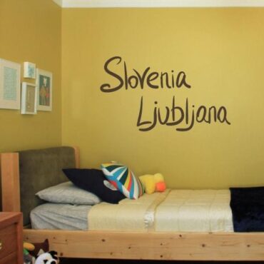 Primer izgleda čokoladno rjave samolepilne stenske nalepke Slovenia Ljubljana pisano na rumeni steni v otroški sobi.