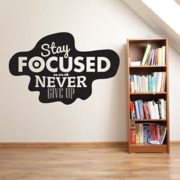 Primer izgleda črne samolepilne stenske nalepke Stay focused and never give up na beli steni v dnevni sobi. Nalepka je napis, ki se glasi: Stay focused and never give up.