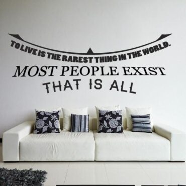 Primer izgleda črne samolepilne stenske nalepke To live is the rarest thing in the world na beli steni v dnevni sobi. Nalepka je napis, ki se glasi: To live is the rarest thing in the world. Most people exist - That is all.