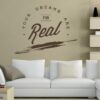 Primer izgleda čokoladno rjave samolepilne stenske nalepke Your dreams are for Real na steni krem barve v dnevni sobi. Nalepka je napis, ki se glasi: Your dreams are for Real.